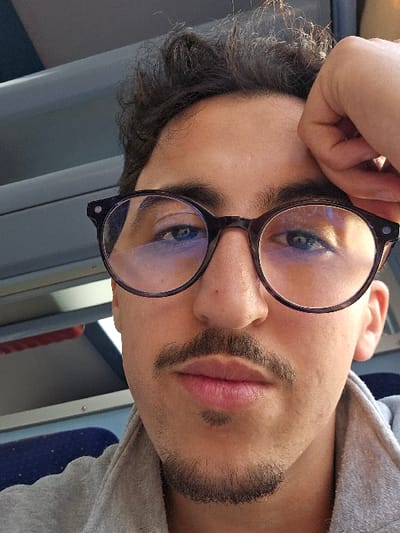A close-up portrait taken in the city of Jbilate الجبيلات, Morocco, features a young adult male with a direct gaze. He has dark, curly hair, dark eyebrows, and visible blue eyes, which are partially obscured by reflections in his dark-rimmed, round glasses. He exhibits a mustache and a chin beard. His right hand is positioned with fingers resting near his forehead on the upper right side of the frame. He is wearing a light grey collared garment. The background elements on the left suggest an interior passenger environment, possibly a train or bus, showing a light grey panel above a section of a dark blue, speckled fabric seat.