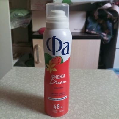 A white aerosol can of "Fa Fiji Dream" deodorant is displayed in the center of the frame, placed on a light-colored, patterned surface. The can features a transparent cap and a prominent "Fa" logo in dark blue. The label depicts an illustration of a slice of guava with a flower and leaf. Below the illustration, text reads "FIJI DREAM" and "DEODORANT." Further down, the can indicates "AROMA OF WATERMELON AND YLANG-YLANG" and "WITHOUT ALUMINUM SALTS." At the bottom, it states "48H PROTECTION AGAINST ODOR" and "150ML." The background is blurred, showing shelves with various items, including what appears to be packaging and possibly some fabrics. The scene is indoors, likely in a domestic setting. There is no indication of its location in Tomilino, Russia.