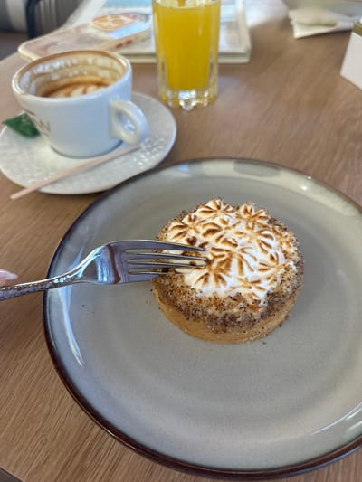An overhead, slightly angled shot shows a breakfast setting on a wooden table. In the foreground, a circular grey plate holds a tart topped with torched meringue, which has a decorative pattern. A fork with a textured handle is in the process of taking a bite from the tart. To the left of the plate, a white cup and saucer hold a partially consumed cappuccino with latte art. A small green leaf rests on the saucer. Behind the cup, a tall glass of orange juice is visible. The background shows a blurred menu or newspaper. The scene is set in Donji Katun, Montenegro.