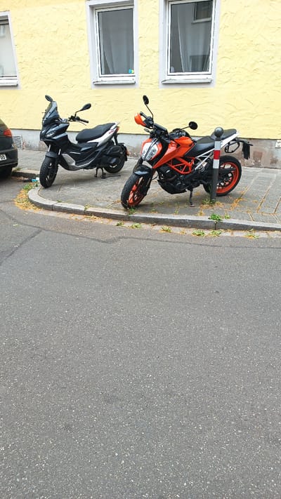 Two parked vehicles are visible on a paved sidewalk in front of a yellow building in Nuremberg, Germany. The vehicle on the left is a black scooter with a windshield. The vehicle on the right is an orange motorcycle with a black frame. Both vehicles are parked in front of a yellow building with two windows. A short, gray, concrete retaining wall separates the sidewalk from the street. The street is asphalt and empty. The image appears to have been taken during daylight.