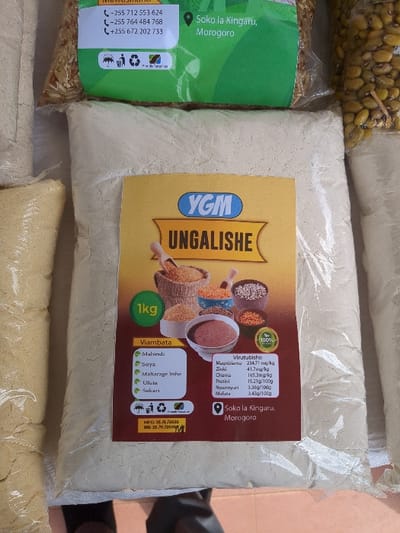 A 1kg bag of Ungalish flour, a brand of blended flour made with mahindi (corn), soya (soy), maharage lishe (nutritious beans), ufuta (sesame seed), sukari (sugar) and other ingredients, is displayed in a shop in Soko la Kingaru, Morogoro, Tanzania, located at latitude -6.829 and longitude 37.658. The bag is sitting on a counter with other bags of flour, rice, and beans behind it. The bag is yellow with black lettering and a red and black label. The label includes nutritional information and a logo for the company. There are no people visible in the image.