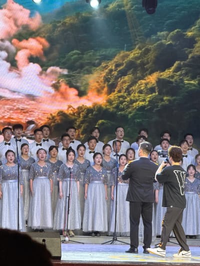 A choir performance is captured on stage, with a large group of singers arranged in rows, facing an audience not fully visible. The image is oriented vertically, but depicts a horizontal scene. The choir members consist of both men and women. The women are dressed in sparkling light blue or silver gowns with sequined bodices and flowing skirts, while the men wear white shirts, black bow ties, and black trousers. All choir members have their mouths open, indicating they are actively singing, and appear focused on their performance.
At the front of the choir, two men are positioned as conductors or directors. The man further back, facing the choir, wears a black long-sleeved shirt with a distinctive white graphic on the back, dark trousers, and white Nike sneakers with a black swoosh. He is holding a smartphone, seemingly recording the performance. In front of him, closer to the choir, stands another man in a black suit jacket and black trousers, also facing the choir, likely conducting.
The stage is equipped with multiple microphones on stands in front of the choir members. A dark, box-shaped piece of audio equipment (possibly a speaker or monitor) is visible on the stage floor to the right. The backdrop behind the choir is a large digital screen displaying an abstract, dynamic image of swirling colors, primarily dark blue, green, orange, and white, resembling clouds, smoke, or a cosmic scene. Bright stage lights are visible on the left side of the frame, illuminating the performers and the backdrop.
The event takes place indoors, likely in an auditorium or performance hall, under theatrical lighting. The scene conveys a sense of concentrated artistic activity during a public performance in Guta, China. There is no discernible text visible in the image.
