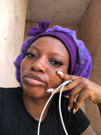 This is a close-up, portrait-style selfie of a young Black woman. She is wearing a purple fuzzy headband with a large bow on the side. She has on a black ribbed shirt and appears to be wearing makeup, including false eyelashes and patterned nail polish. Her right hand is raised, with her index finger touching her cheek, and white earbuds are visible. The image is oriented sideways.
The background suggests an outdoor or semi-outdoor setting, possibly an alcove or underpass, with textured concrete walls and a beige, possibly tiled, surface. The lighting appears to be natural and somewhat diffuse, hinting at daytime. There are no discernible signs of weather conditions. The woman's expression is neutral, and she is looking towards the camera. The overall impression is a casual, self-taken photograph.