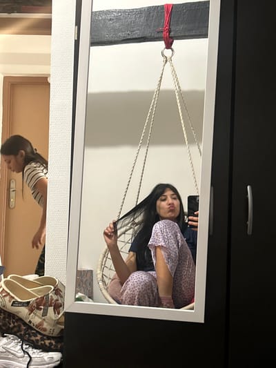 A girl in pink pajamas sits in a hanging chair visible in a mirror's reflection. She's taking a selfie, holding her phone in one hand while tugging on her hair with the other. The mirror is hung by rope tied to a support beam with red material. The reflection below the mirror shows another part of the room with a door and shoes on the floor. A dark dresser with a drawer pull frames the top of the mirror.