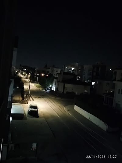 This image presents an elevated view of a quiet urban street at night, likely in North Nicosia, Cyprus. The scene is dominated by a curving, paved road, unevenly illuminated by several streetlights that cast bright pools of light and deep shadows across the asphalt.
On the left side of the street, a light-colored sedan is parked directly under a prominent streetlight, its surface reflecting the artificial light and creating a stark silhouette. Residential buildings, ranging from two to several stories, flank both sides of the road. Many windows in these buildings show interior lights, suggesting occupancy. On the right, a long, low white wall runs parallel to the street.
The road surface exhibits several parallel linear markings or grooves, stretching into the distance. The sky above is dark and clear, indicating a cloudless night. In the bottom left corner, some indistinct items are visible hanging from what appears to be a balcony or railing, including what looks like a pair of gloves.
The image conveys a sense of stillness and quiet urban life after dusk. No people or active events are captured. A digital timestamp in the lower right corner reads "22/11/2025 18:27," confirming the time of day as early evening.