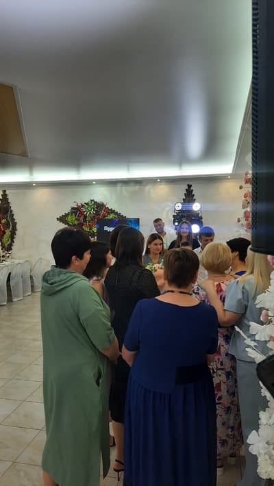 The image depicts a gathering of people inside a dimly lit event hall. Several individuals, mostly women, are seen from behind, facing a group of people in the center of the room. The background features a white wall with decorative elements and lighting. The floor is tiled with a light-colored pattern. The overall scene suggests a celebratory or social event. The location is specified as within the Жодзішкаўскі сельскі Савет, Belarus.