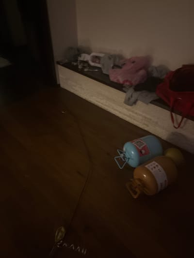 The image depicts an interior scene, likely a room, with a wooden floor and a white, built-in shelf or counter along the wall. On the shelf, several items of clothing are piled, including a pink garment that appears to be a stuffed toy or bag, and some gray clothing. To the right of the clothing, a red bag is visible. On the floor in the foreground, there are two cylindrical objects, possibly canisters or tanks, one blue and one orange. The blue object has a handle and labels with text and a QR code. The orange object also has labels. The room is dimly lit, creating shadows and a somewhat cluttered appearance. The location is stated to be in the city of Orekhovka, Russia, but there are no specific visual cues in the image to confirm this geographical location. There are no people visible in the image, nor any actions or interactions taking place.