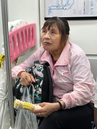 A middle-aged East Asian woman sits on a subway train, holding a partially eaten corn cob in her right hand. She is wearing a pink zip-up jacket with a white tag that reads "PaiLaiYa". The woman has dark hair and appears to be looking upwards and to her left with a neutral expression. In her lap, she has a backpack with a camouflage pattern. A clear plastic bag containing the corn cob is also visible. Behind her, a pink, curved rack is mounted on the wall, and a white garment with a zipper is partially visible on the left. A subway map is displayed on the wall above and behind her. The scene appears to be in Longta, China.