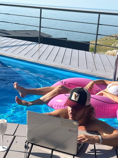 A person is working on a laptop poolside. They are wearing a black baseball cap with a patch of an eagle on it and a maroon brim. The person is sitting on the edge of a pool with their legs dangling in the water. Beside them is a pink inflatable flamingo, and another person is seated on it. The pool is surrounded by a wooden deck. In the background, there is a body of water, likely the sea, and a grassy hillside. The sun is shining, casting shadows on the deck, suggesting it is daytime. The overall scene depicts a relaxed, yet productive atmosphere.
There is a white bracelet on the person's wrist with some text that is partially visible, but not legible. A white wine glass is also visible on the deck. The location context provided is Мысовское сельское поселение, Ukraine.