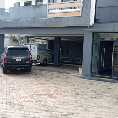 The image depicts a paved driveway in front of a modern building with a dark gray facade and a glass entrance. Two vehicles are parked on the driveway: a dark-colored Toyota 4Runner SUV in the foreground and a white pickup truck with a utility box in the background. The SUV has the license plate "MUS 942 JB". Palm trees and other greenery are visible behind the vehicles, suggesting a tropical or subtropical climate. The building features large windows and multiple levels, with a balcony or overhang on the second floor. The sky is not visible, but the lighting suggests it is daytime. The scene appears to be a private or commercial property in Ibadan, Nigeria. No people are visible, and the overall atmosphere is still and quiet. The paving stones are laid in a herringbone pattern. Visible text includes "TOYOTA" and "4RUNNER" on the SUV, and lettering on the utility box of the white truck which includes a phone number "08140666666".