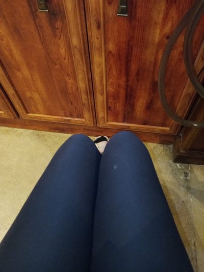 The image depicts a first-person, downward-facing view of a seated individual's legs in an indoor environment within Madinaty, Egypt. The subject's legs are clad in dark blue form-fitting leggings, with a small white mark visible on the right leg. The tip of a light-colored sneaker, featuring a white sole and a beige or pink upper, is visible between the knees. Directly in front of the legs are two reddish-brown wooden cabinet doors, each equipped with a dark metallic handle. The wood exhibits distinct grain patterns. To the far right, a dark, curved hose or cable is partially visible against the cabinet. The floor beneath the legs and cabinets consists of light-colored tiles or stone with visible grout lines and minor imperfections.