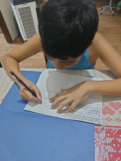 A young boy is focused on drawing on a piece of paper laid out on a blue folder. He is wearing a blue tank top and has dark, short hair. The paper has a comic strip layout with boxes for drawing and lines for text. The boy is holding a black pen and appears to be diligently working on his drawing and writing.
In the background, a white and grey air cooler is visible, suggesting an indoor setting. The table is covered with a patterned cloth featuring small red hearts.
On the paper, there is some handwritten text in what appears to be Filipino. One section reads "AKIN ANG LUPANG ITO," which translates to "This land is mine." Another section contains more text, possibly a story or caption for the comic strip. The OCR also detects text that may be part of a larger narrative related to land disputes in Mindanao.
The overall scene depicts a child engaged in an educational or creative activity, possibly a school assignment, at home. The lighting suggests it is daytime.