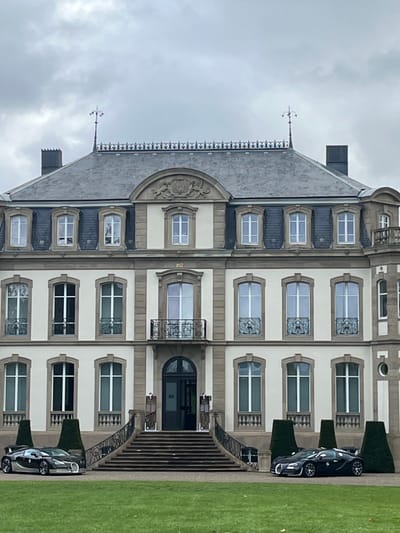 The image displays a frontal view of the Château Saint-Jean in Dorlisheim, France, under an overcast sky. The elegant, three-story building features a light-colored facade with darker stone accents around windows and corners, and a steep grey slate roof adorned with dormer windows, ornate ironwork along the ridge, and decorative finials. A grand central staircase with dark railings leads to the main entrance, which has a dark wooden door. Two Bugatti Veyron supercars are parked symmetrically on paved areas on either side of the staircase. The vehicle on the left is a two-tone silver and dark grey/black, while the vehicle on the right is solid dark grey/black. Both cars face forward. Manicured conical hedges are positioned near the vehicles and the staircase. The foreground is a well-maintained green lawn. No individuals are present in the scene.