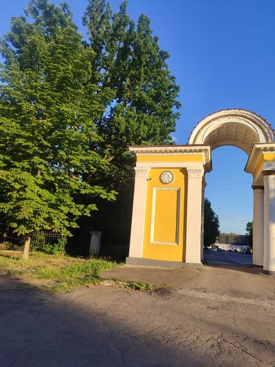 A bright yellow and white arched gateway stands on a cracked asphalt path. The arch is ornate, with detailed white molding and a terracotta-colored edge. On the left pillar of the gateway, a round clock face is embedded within a decorative white circle. Behind the gateway, a building with a glass facade is visible, with several cars parked in front of it. To the left of the gateway, lush green trees with full canopies dominate the scene, their leaves illuminated by sunlight. A metal fence runs along the edge of the property. The sky above is a clear, vibrant blue. The scene is located in the city of Zhovti Vody, Ukraine.