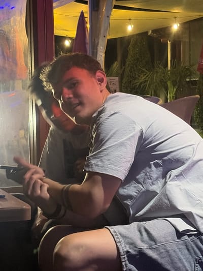 This image, captured in Elhovo, Bulgaria, depicts a nocturnal scene on what appears to be an outdoor patio or restaurant area. The primary subject, a young male, is seated on the right side of the frame, facing partially towards the viewer. He possesses dark, curly hair and is wearing a light grey short-sleeved t-shirt and blue denim shorts. His right arm is bent, and his hands are interlocked or holding a dark, rectangular object, possibly a smartphone, near a light brown wooden table surface. A dark band is visible on his right forearm. He is looking directly at the camera with a slight expression. Behind the primary subject, a second male individual is partially visible, blurred and in shadow, wearing a light-colored shirt, with only his head and shoulder discernible. The background features a stretched yellow or beige awning illuminated by multiple string lights with exposed bulbs, indicating an evening or nighttime setting. Dark green foliage, consistent with ornamental plants or bushes, is visible behind the individuals, alongside indistinct building structures or pillars. The overall environment suggests a casual, artificially lit outdoor setting.