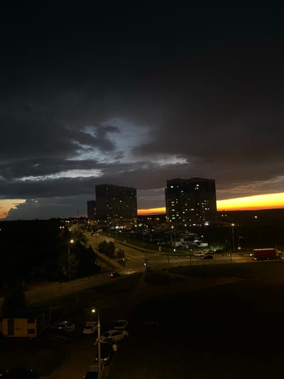 The image depicts a twilight scene in Domodedovo, Russia. The sky is a dramatic blend of dark clouds on the left, gradually transitioning to a vibrant orange and yellow band near the horizon on the right, suggesting sunset or sunrise. Silhouetted against the colorful sky are two multi-story apartment buildings with numerous illuminated windows, indicating that it is evening and people are home.
Below the buildings, a road is visible with streetlights casting a warm glow. Several parked cars are scattered along the roadside and in a parking lot. A large red truck is parked near the top right of the frame. The overall setting appears to be a residential urban or suburban area. The time of day is clearly either dusk or dawn, characterized by the dramatic sky colors and the lit windows in the buildings. The weather appears to be transitioning, with dark, possibly stormy, clouds present alongside the clear, bright horizon. No people are clearly discernible in the image. No visible text is present.