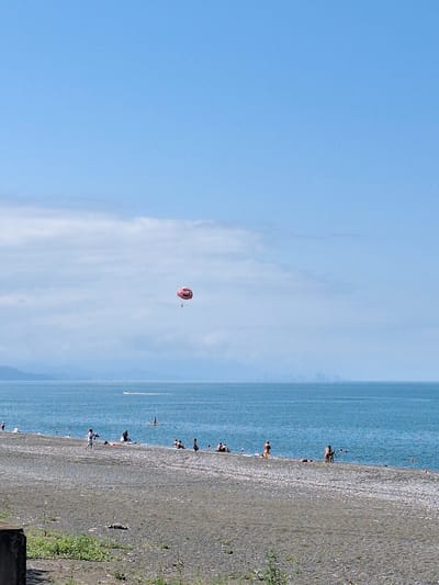 The image captures a serene beach scene in Kobuleti, Georgia, on a clear, sunny day with a bright blue sky and a few wispy clouds. The vast expanse of the Black Sea dominates the upper half of the frame, its surface a deep, calm blue, with a faint, distant shoreline visible. A single parasailer, suspended from a pink parachute, is a small, vibrant detail in the sky.

The foreground is a wide, pebble-strewn beach. Several people are scattered across the sand, some sunbathing, others walking or standing near the water's edge. A few individuals are visible in the shallow sea. In the distance, a small boat leaves a white wake on the water, and further out, the faint silhouette of a city or town can be seen on the horizon. The overall atmosphere is one of leisurely summer recreation by the sea.