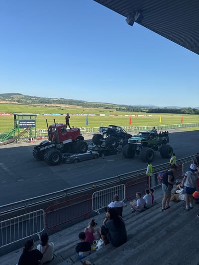 The image captures a sunny day event at Punchestown Racecourse, Ireland, featuring monster trucks and spectators. In the foreground, a group of people are seated on concrete bleachers, facing away from the camera, observing the event. Beyond them, a barrier separates the spectator area from the event space where several large monster trucks are positioned. One prominent red monster truck, with a driver standing on its roof, is situated in the center. To its right, a green monster truck is also present, alongside a black monster truck. Several crushed cars are visible beneath the trucks, suggesting a stunt performance. In the background, the expansive green fields of the racecourse are visible, bordered by trees and rolling hills under a clear blue sky. Various advertisements and banners are displayed along the racecourse perimeter. Two individuals wearing high-visibility vests are seen near the trucks, possibly event staff.