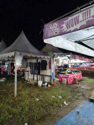 The image depicts an outdoor night market scene in An Khe Ward, Vietnam, illuminated by artificial lighting under a dark sky. The foreground shows uneven, muddy ground with sparse green grass and scattered litter, including plastic bags. Several white or grey conical canopy tents are erected across the area. In the mid-ground, one tent displays clothing items, such as shirts and towels, hanging from its interior structure. Near this tent's base, multiple buckets (white, red, yellow) and metal containers are present. To the right, a large white canopy features a purple banner with "GO CENG SO" and "WOW...!!!!" printed on it. Under an adjacent canopy, a market stall is visible with a pink and white banner displaying text including "M.J.", "KOLLAGEN", and "KL", with various items arranged on a table. A small wooden stool sits beside this stall. Further in the background, additional market stalls are visible with bright internal lighting and various goods. A blue tarp lies partially on the ground in the lower right. No individuals are clearly visible.