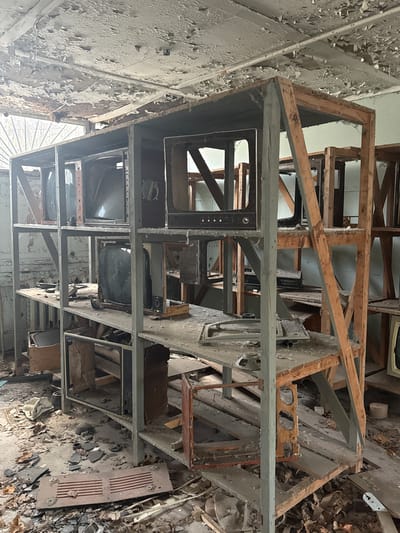 The image depicts a collection of old television sets stacked on a wooden and metal framework inside a dilapidated building. The walls are heavily peeling, revealing layers of paint and structural decay. The floor is covered in debris, suggesting a state of abandonment and neglect. The location is likely in the city of Pryp'yat', Ukraine, based on the context of abandoned buildings often associated with the Chernobyl Exclusion Zone. There are no people visible in the image. The primary subjects are the decaying televisions and the crumbling state of the building's interior. The interaction is of objects with their environment, undergoing decay and disintegration.