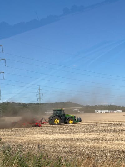 In the city of Telnice, Czechia, a green and yellow John Deere tractor is actively plowing a dry, harvested field under a clear blue sky. The tractor is in motion, kicking up a significant amount of dust from the tilling process. Behind the tractor, a red plowing implement is attached, digging into the soil. In the background, power lines stretch across the frame, supported by tall metal pylons, and a line of trees and hills with some buildings are visible in the distance. The field in the foreground is covered in stubble from a previous crop, with some small purple flowers scattered among the dry vegetation. The overall scene depicts agricultural work taking place on a sunny day.