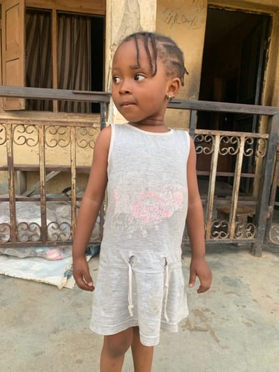 A young, dark-skinned girl stands in front of an old, weathered building with decorative wrought iron railings. She wears a light grey dress with a faded design and has braided hair. Her expression is curious, as she looks off to the side. The background features a combination of textured walls, dark doorways, and a glimpse of a white material on the ground. The image appears to be taken outdoors in daylight, with a slight tilt.