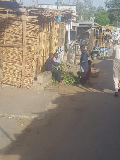 A daytime street scene in Dabawa, Nigeria, shows two individuals sitting on a concrete structure next to a hut-like building constructed of thin bamboo-like poles. One person is seated while the other appears to be either lying down or leaning back. Another individual stands nearby, and further down the road, a group of people are gathered around what appears to be market stalls or makeshift shops constructed of wood. The street surface is paved and appears to be well-used. A variety of objects are also scattered around the ground, including leaves and discarded items.