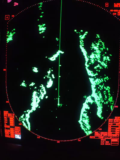 A radar display screen, likely from a marine vessel, is shown against a black background, illuminated with bright green radar returns and red text and numerical readouts. The main subject is the circular radar scope, which features scattered and dense green shapes indicating landmasses or other significant objects.

A prominent vertical green line extends from the center towards the top, likely representing the vessel's heading or a bearing line. The display includes red radial markings for bearings, with numbers such as "330," "300," "270," "240," "210," "180," "150," "120," and "090" visible around the perimeter.

In the upper left, "SP" and "2km" indicate the range scale. Additional numerical data, including "2.00S," "88-888-," and "030," appear in the upper right. Red graphical icons, such as a square with a triangle and another red square, are also present.

On the lower left and right portions of the screen, various radar settings and display options are shown in red text. These include:
*   "DISP STD BRILL 1" with sub-options like "EBL," "VRM," "NAV," "HL," "RINGS," "08-TK," "MARK," "DATA," "TRAIL," "PANEL BRILL," "ECHO COLOR," and "BACK CO(LOR)".
*   "DISP STD ECHO 1" with settings such as "FTC OFF," "IR," "ES OFF," "NR OFF," "REL 2SCAN EAV," "TRAILS OFF," "CONTRAST 1," "AUTO TUNE," "GAIN," "A/C SEA," and "A/C RAIN."

The green radar echoes suggest navigation in an area with irregular landforms, possibly a coastline or islands. No people are visible, and the time of day is not discernible from the image, though the presence of "A/C SEA" and "A/C RAIN" settings indicates the system's ability to operate in various weather conditions. The overall scene depicts a functional nautical navigation interface. The location context provided is Vorob'evo, Russia.