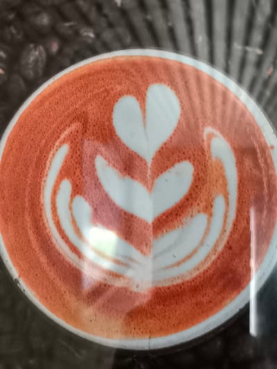A close-up overhead shot shows a coffee with latte art, likely in Vulcănești, Moldova. The coffee is in a white cup, with a circular orange-colored foam topping. The foam has been artfully arranged to create a pattern resembling stylized leaves or petals, with a central heart shape at the top. The background is dark and out of focus, suggesting an indoor setting like a cafe or kitchen. The lighting creates some glare on the surface of the coffee.
