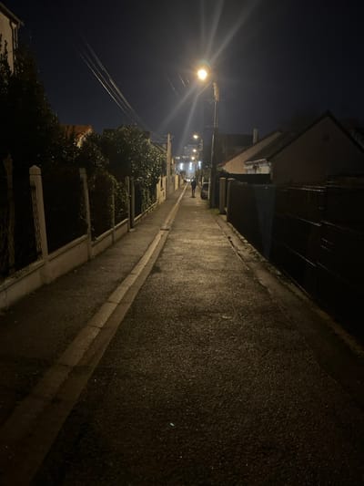 A narrow street at night stretches into the distance, illuminated by distant streetlights. Buildings line both sides of the road. On one side, foliage peeks over what appears to be a wall. The road's surface glistens, reflecting the dim light. A lone figure walks in the distance. Electrical wires cut across the dark sky above.