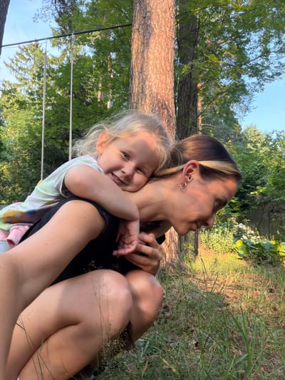 In this candid outdoor photograph, a woman and a young child are embracing, seated amidst a natural setting. The woman, with blonde-streaked brown hair and a hoop earring, looks upwards and to the right with a gentle expression. The child, with blonde hair, is nestled in the woman's arms, smiling broadly at the camera and hugging the woman's neck. Both are dressed casually; the child wears a tie-dye shirt, and the woman a black top.
The background suggests a park or wooded area in Udelnaya, Russia. A large tree trunk, with rough bark, is prominent to the left, and lush green foliage and trees form the backdrop. The scene appears to be lit by natural sunlight, indicating it's likely daytime, possibly late afternoon given the warm, golden quality of the light filtering through the trees and illuminating the grass. The ground is covered with dry grass and some scattered dry seed heads. In the upper left quadrant of the image, a portion of what appears to be a hammock or swing set with ropes and metal supports is visible against a clear blue sky. The overall mood is one of warmth, affection, and peaceful enjoyment of nature. There is no visible text in the image.