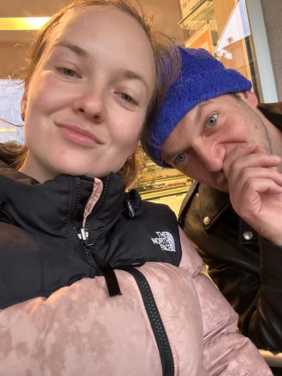 A selfie-style photograph depicts two individuals within a commercial establishment in Longone al Segrino, Italy. The primary subject, an adult female positioned on the left, has light skin and light brown hair. She wears a black "THE NORTH FACE" branded puffer jacket over a light pink garment and displays a subtle smile while looking directly at the camera. To her right, partially obscured, is an adult male with blue eyes and facial stubble, wearing a bright blue knit beanie and a black leather jacket with visible metal snaps. His right hand is raised to his face, partially covering his mouth and nose, as he also looks towards the camera. The background, intentionally blurred, reveals a glass display case filled with baked goods or pastries, indicating an interior setting consistent with a cafe or bakery. Reflections in background glass suggest an exterior urban environment.