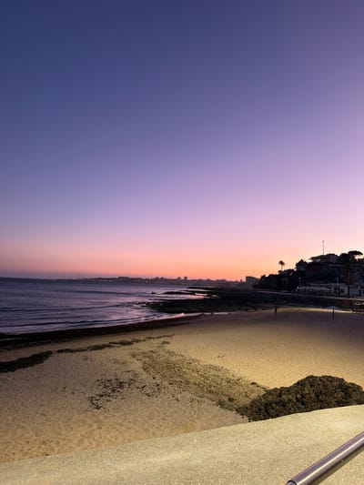 An image captures a panoramic view of a beach and coastline in Cascais, Portugal, during twilight. The sky transitions from a deep purple at the top to shades of pink and orange closer to the horizon, where a cityscape with buildings and cranes is visible in the distance. The sea is calm with gentle waves lapping against the shore. The beach is sandy, with scattered patches of seaweed and a large pile of dark material, possibly more seaweed or debris, towards the foreground. A paved walkway runs along the edge of the beach, illuminated by streetlights. A lone figure is seen walking on the beach in the middle ground. The foreground features a concrete barrier with a metal railing.