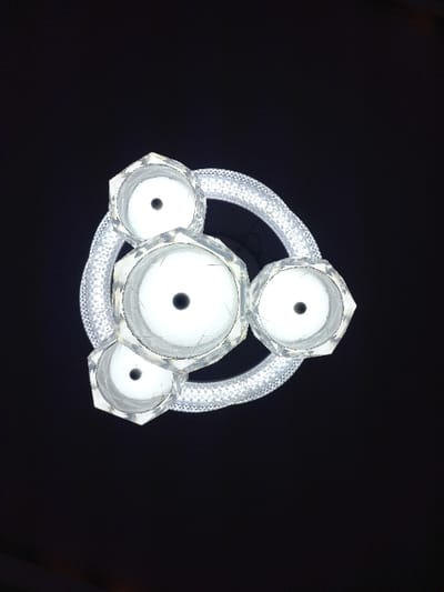 A circular, sparkling light fixture dominates a black background. It consists of four faceted light bases arranged around a ring of smaller, sparkling elements. Each base appears hollow, with a dark center. The central base is larger than the others, with intricate facets. The ring encircles the structure, connecting to each light base. The fixture emanates a bright, white light.