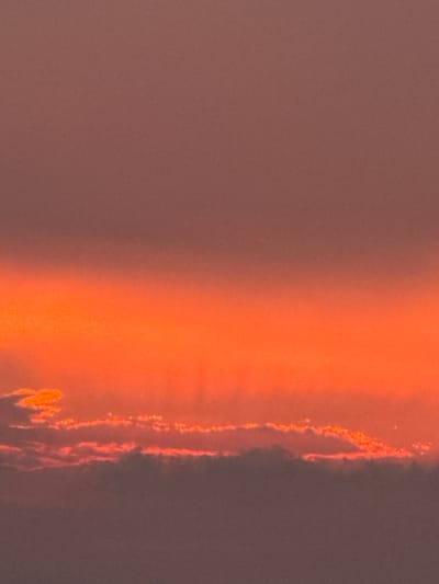 The image depicts a sunset over the city of Surgut, Russia. The sky is filled with a gradient of colors, transitioning from a deep reddish-brown at the top to a vibrant orange and yellow towards the horizon. Wispy clouds are visible, illuminated by the setting sun, with some appearing tinged with pink and orange. The lower portion of the image is darker, suggesting the ground or a distant landscape. No people or specific objects are identifiable, creating an abstract and atmospheric scene focused on the natural beauty of the sunset. The image is a photograph with a textured quality, possibly due to the camera's processing or the lighting conditions.