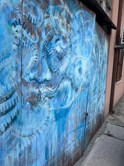 The image shows a close-up of a rusted metal surface painted with abstract blue and white swirls. The surface appears to be a garage door or a similar external structure. The paint is applied in thick, gestural strokes, with some areas showing the underlying rust and metal texture. There are vertical lines suggesting the surface is made of planks or panels. A lock is visible on the right side of the door, and a small padlock is attached to it. To the right of the painted surface, a section of a pink-colored building is visible, with a brown shuttered window and a downspout. A concrete sidewalk runs alongside the building and the painted surface. The scene is located in the city of Cetinje, Montenegro.