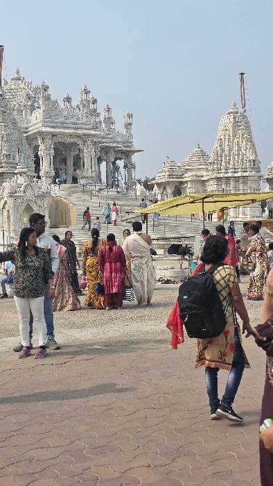 This image captures a vibrant scene at a temple complex, likely the Shri Digambar Jain Siddh Kshetra Parasnath Atishay Temple in Madhuban, India, given the location context. The dominant feature is a large, ornate white stone temple structure with multiple towering spires (shikhara) and intricate carvings, characteristic of Jain architecture. The temple appears to be made of white marble or a similar light-colored stone, gleaming under bright sunlight.

Numerous people are present, moving and standing on the paved ground surrounding the temple. Most individuals appear to be adults, with some possibly being families. They are dressed in a mix of traditional Indian attire, such as sarees and kurtas, and casual Western wear like jeans and t-shirts. Many are seen walking in groups or individually, suggesting they are visitors or devotees. Some individuals are carrying backpacks or handbags. A large yellow umbrella or awning provides shade in one area near the temple steps.

The scene takes place during the daytime under a clear, bright sky, indicated by the strong sunlight and visible shadows cast on the ground. The weather appears fair and sunny. The ground is paved with interlocking tiles featuring a distinctive wavy pattern. There is no clearly decipherable text visible in the image. The overall atmosphere suggests a busy religious or tourist site, with people engaged in their visit to the sacred grounds.