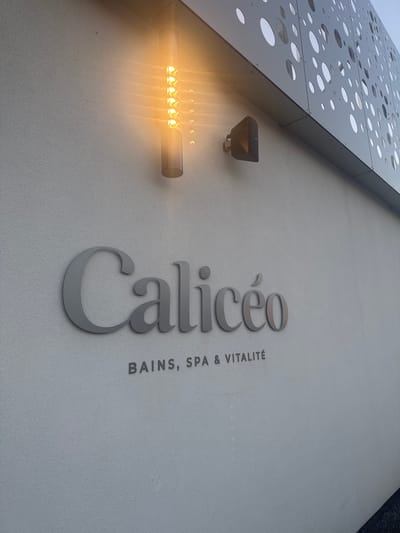 The image depicts a building facade in Marcq-en-Barœul, France. The building features a modern design, with a perforated metal panel at the top-left corner. Below, on a light-colored wall, the name "Calicéo" is displayed vertically in stylized, gray lettering. To the right of "Calicéo" is the phrase "BAINS, SPA & VITALITÉ" in smaller, sans-serif letters. To the left of the logo are two light fixtures, one rectangular with several small bulbs emitting a warm light, and a smaller, box-shaped fixture above it. The overall scene suggests a spa or wellness center.