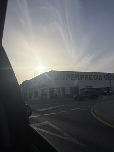 This is a medium-shot, eye-level photograph taken from inside a vehicle, looking out at a street scene in Urbanización La Cenia II, Spain. The sun is low in the sky, creating a bright glare and casting long shadows. A large building with the words "SUPERPRECIO CHINO" emblazoned across its facade dominates the background. Several cars are visible on the road in front of the building, including a dark SUV. The road surface is asphalt with visible white lane markings and a yellow curb. The sky is partly cloudy with wispy formations. The overall lighting suggests it is either early morning or late afternoon.