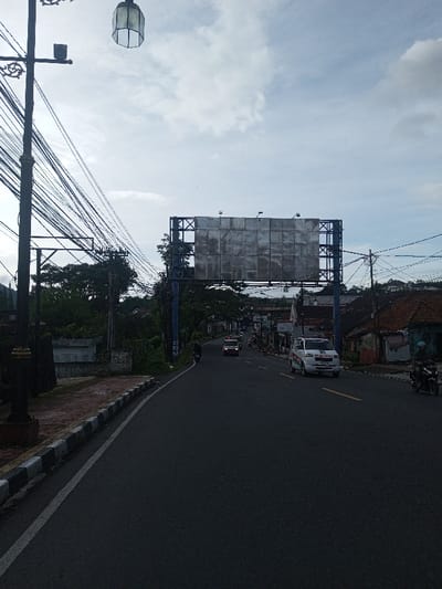 A road stretches forward beneath an overcast sky. Utility poles laden with wires line the left side of the road. A large, blank billboard spans the road ahead. Vehicles, including an ambulance, are visible driving in the distance. A curb runs along the left edge of the road. A streetlamp stands tall in the left foreground.