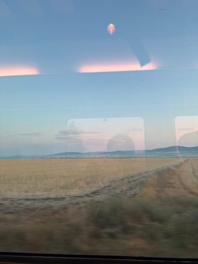 The image shows a view from inside a vehicle, looking out a window at a landscape. The sky is a pale blue with soft pinkish-orange clouds near the horizon. A faint reflection of what appears to be interior lights and a small, abstract red and yellow object are visible on the window. Below the sky, a vast field of dry, golden grass or crops stretches towards distant, hazy blue hills. A dirt road or track cuts through the field diagonally from the right side of the frame, leading towards the hills. There are also indistinct reflections of the vehicle's interior on the window, including rectangular shapes that suggest other windows or panels. The overall impression is of a rural landscape viewed during twilight or dawn. There are no people or discernible activity in the scene. The provided information about Chernograd, Bulgaria, does not directly correlate with any visual elements in the image itself.