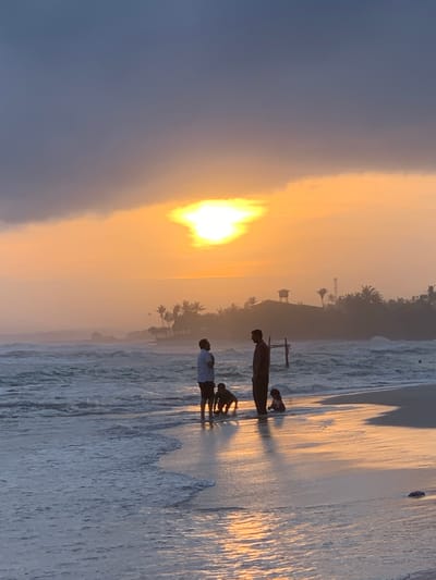 This image depicts a serene sunset scene on the beach in Kathaluwa, Sri Lanka. In the foreground, the gentle waves of the ocean lap the shore, reflecting the warm hues of the setting sun. Three individuals and two dogs are silhouetted against the vibrant sky. Two adults, one standing and the other bent over, are interacting with two young children who are playing near the water's edge. The backdrop features a hazy coastline dotted with palm trees, and a building with a water tower. The sky is a canvas of dramatic colors, with dark clouds parting to reveal a brilliant sun, casting a golden glow over the entire scene. The overall atmosphere is one of tranquility and family enjoyment by the sea.