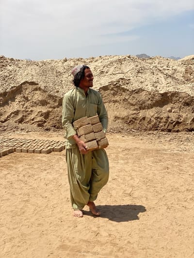 In Deh-E Bagh, Afghanistan, a barefoot man is carrying a stack of bricks in front of a large pile of earth, with rows of drying bricks laid out in the background. He is wearing loose-fitting green clothing and a cap.