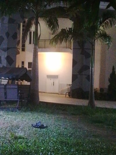 A nighttime outdoor scene features a building facade with palm trees. The building has geometric patterns, windows, and a balcony. A bright light shines from within a recessed area with double doors. In the foreground, a bench swing set sits on a lawn with what appears to be a pile of debris. The scene's dark lighting adds to the mystery of the setting.