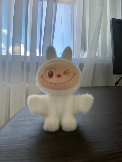 A white, fluffy plush toy with large, round ears and a smiling face with one eye winking and a red nose is positioned in the foreground of the image. The toy is sitting on a dark, textured surface, likely a desk. Behind the toy, sheer white curtains are drawn across a window, revealing a bright blue sky with some clouds visible. To the right of the toy, partially visible, is a black computer monitor. The lighting suggests it is daytime. The image was taken in Uchaly, Russia.