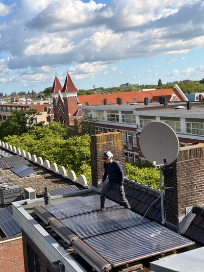 The image shows a rooftop scene in Rotterdam, Netherlands. The main subject is a person wearing dark clothing and a white and black patterned hat, who is standing on a rooftop covered in solar panels. The person appears to be working on or near the solar panels, with their hands positioned as if they are making an adjustment.
The rooftop is multi-leveled and features a variety of elements. There are numerous solar panels and what appear to be solar water heaters. A large satellite dish is also prominently visible. The surrounding buildings are residential, with red-tiled roofs and brick facades. Green trees are interspersed between the buildings, indicating a somewhat urban but leafy environment.
The sky is partly cloudy, with large, dramatic cloud formations suggesting it is either morning or late afternoon. The lighting indicates it is daytime. The overall atmosphere is one of a task being performed outdoors on a bright but not entirely clear day. There is no visible text in the image. The perspective is from a higher vantage point, looking down and across the rooftops.