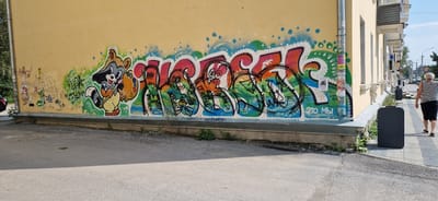 The image shows a section of a building with graffiti on its exterior wall, located in the city of Miass, Russia. The graffiti features a cartoon raccoon holding a palette and paintbrush, alongside abstract, colorful lettering and designs. The building itself is a light yellow, with architectural details such as windows and decorative moldings visible. A sidewalk runs alongside the building, and a portion of a street is visible in the foreground. A person, a woman with white hair wearing a striped tank top and black shorts, is walking away from the camera on the sidewalk. There are several dark-colored trash receptacles placed along the sidewalk. The overall scene depicts an urban environment with street art.