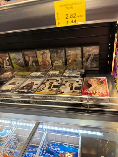 The image shows a refrigerated display case in a store in Pravets, Bulgaria. The case contains various frozen food items, including ice cream and bagged snacks. Above the refrigerated display, on a shelf, there are several audio cassettes for sale. A yellow price tag is visible at the top, indicating a price of 2.82 BGN and a sale price of 1.44 BGN for "OSHEE PORTOCALI 750 ML". The cassettes are packaged in plastic cases, and some of the covers feature images of people, musical instruments, and landscapes.