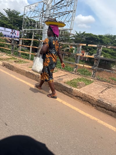 A person walks on a paved road with a yellow line marking the lane. The person is wearing a dark dress with an orange and black floral pattern, a pink headscarf, and yellow footwear. They are carrying a white bag in their left hand. On their head, they balance a yellow round object, possibly a basin or hat, with dark round items inside, likely fruits or vegetables. To the left of the road, there is a raised concrete walkway bordered by a rusty metal fence. Beyond the fence, lush green trees and vegetation are visible, along with some buildings. A large white metal structure, possibly a billboard frame, stands in the background, with a banner displaying "MADUKA UNIVERSITY" and images of students. The sky is partly cloudy, suggesting daytime. The scene appears to be a street in Enugu, Nigeria. The lighting and shadows indicate it is likely midday or early afternoon. The person's posture suggests they are walking with purpose, carrying their goods.