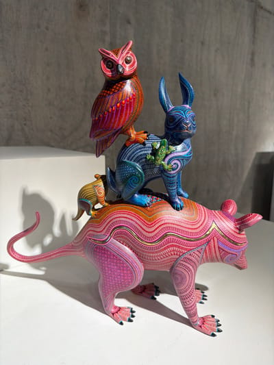 The image displays a vibrant stack of three intricately carved and painted wooden animal figures, characteristic of Mexican *alebrijes* from San Martín Tilcajete. These fantastical creatures appear to be climbing or clinging to each other and a plain white wall, creating a dynamic, vertical composition.
The largest figure, a pinkish-red creature resembling a bear or a stylized monster, clings to the white wall on the right. Its body is covered in elaborate, swirling patterns of various shades of pink, red, orange, and gold, with distinctive black-tipped claws. Positioned on the back of this pink creature is a smaller, blue animal, possibly a rabbit or a similar mythical beast, adorned with complex, wave-like patterns in blues, purples, and greens. A small, bright green frog or lizard with dark spots clings to the blue creature's back, near its head. The third figure, an orange and red owl with its mouth slightly ajar, perches atop the blue creature, showcasing detailed feather-like designs and intricate patterns in warm hues.
The bright, directional lighting from the left casts prominent shadows on the white wall and the neutral grey background, emphasizing the three-dimensionality and playful interaction of the sculptures. The scene takes place indoors, likely in a gallery or art display setting, highlighting the meticulous craftsmanship and vibrant colors associated with traditional Mexican folk art. No text is visible in the image.